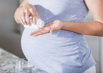 Cropped view of pregnant woman taking pill with glass of water, possibly a prenatal vitamin.