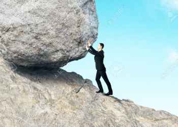 Side view of young businessman pushing huge rock up mountain. Beautiful background. Hard work concept