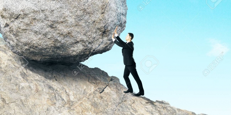 Side view of young businessman pushing huge rock up mountain. Beautiful background. Hard work concept